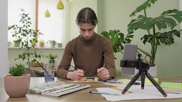 Teenage Guy Drawing with Watercolors Watching Video Class on Smartphone alt
