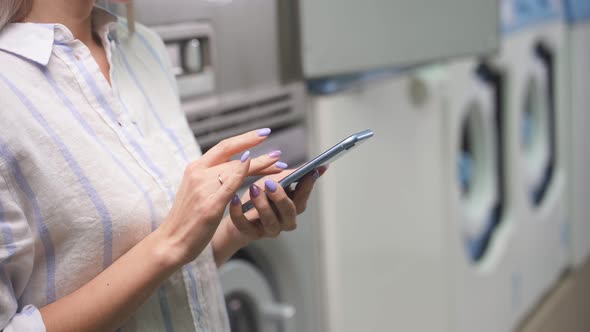 A Woman in a Public Laundry Holds a Smartphone in Her Hand and Conducts Correspondence While Washing