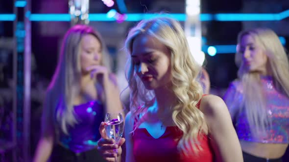 Relaxed Blond Woman with Wine Glass in Red Dress Dancing in Night Club Lights with Blurred People at alt