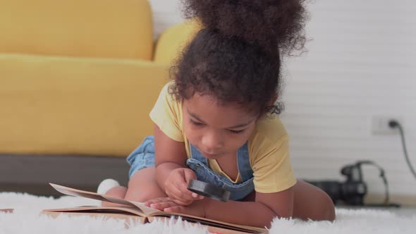 Little kid girl reading book with magnifying glass on the floor in living room