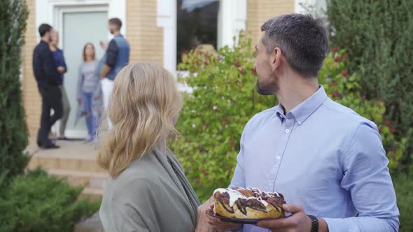 Happy Mature Couple with Cake Smiling and Looking at Camera Standing in the Foreground While Company alt