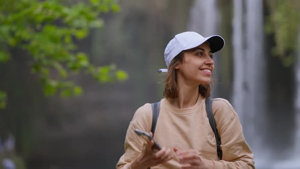 Close Up of Beautiful Young Smiling Woman Tourist Taking Photo or Video on Mobile Phone on Waterfall alt