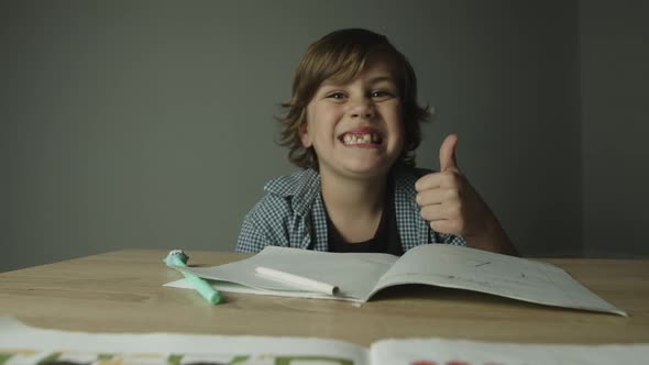 Cute Child Schoolboy Sits at Desk with Notebooks and Pencil Finishes Homework Shows Thumbs Up and
