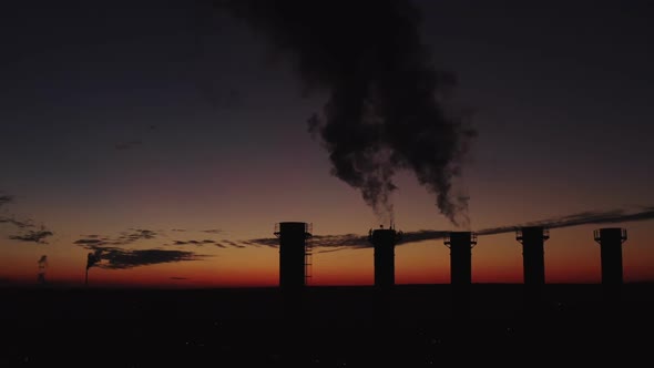 High Chimneys Against the Backdrop of the Sunset and the City. Thick Smoke Rises High. Silhouette of alt