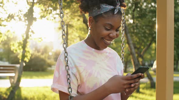 Joyous African Woman Sitting on Swing in Park and Using Phone alt