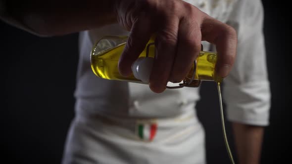 Italian Chef in the Kitchen in a White Robe Pours Yellow Olive Oil Over a Dish.