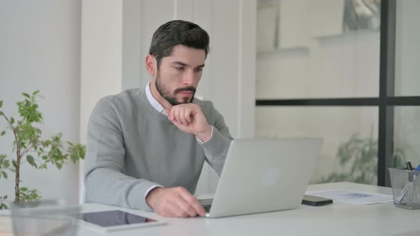Young Man Thinking While Working on Laptop in Office alt