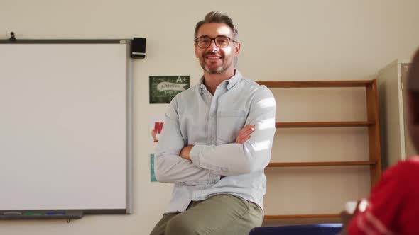 Portrait of happy caucasian male teacher sitting on desk in classroom looking at camera alt