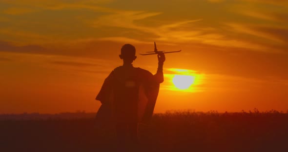 A Boy Is Going Across a Field with a Plane in His Hand. Sunset.  alt