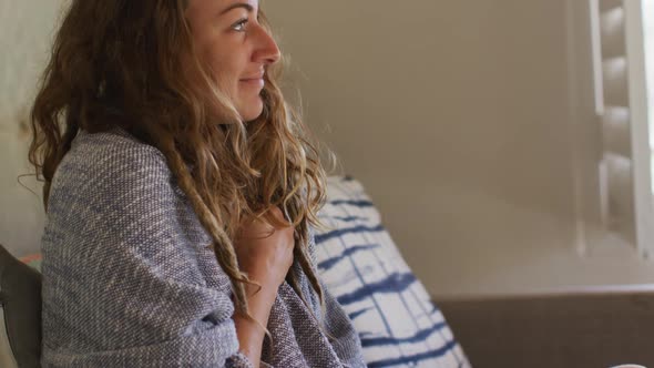 Caucasian woman sitting on sofa with blanket around shoulders smiling in sunny cottage living room alt