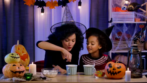 Mom and Daughter African American in Festive Costume and Witch Hat Having Fun and Drinking Hot alt