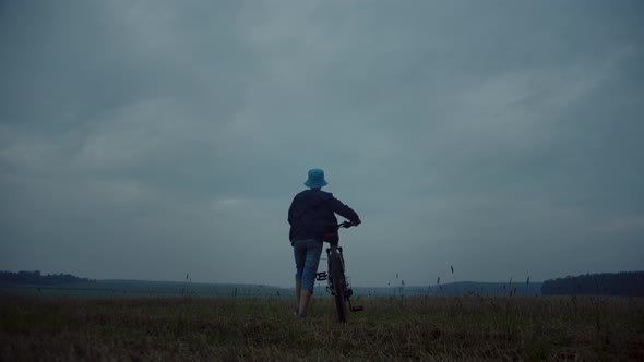 Boy With A Bicycle In A Field In Rainy Weather Looks Around at the Top of the Field Goes Into the