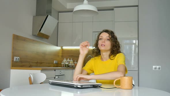 Tired Young Woman with Curly Hair and Yellow Shirt is Working From Home Using Her Laptop at the alt