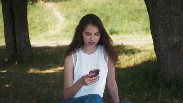 Beautiful Cheerful Girl with a Smartphone Sitting in a Park on a Bench on a Sunny Day Teenager alt