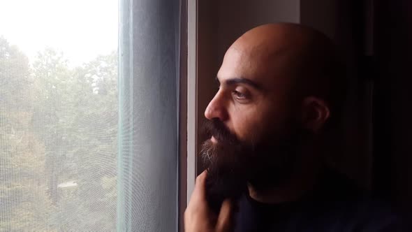 Young bearded caucasian man indoor looking outside window touching beard laughing alt