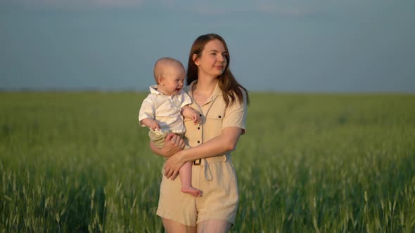 Mom with a Ten Month Old Son in Her Arms Walks Through a Green Wheat Field and Enjoys the Warm Sunny