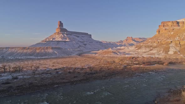 Flying up and over trees past river towards desert cliff spire in winter alt
