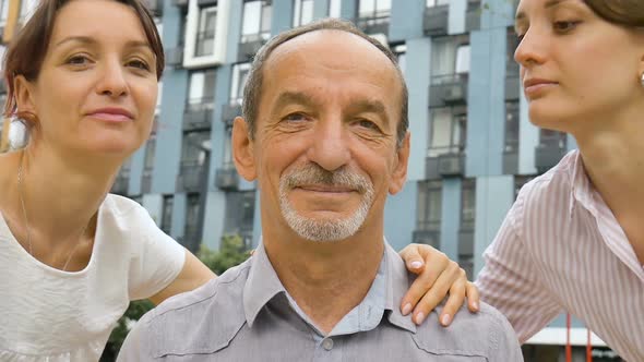 Family Portrait of Senior Father and Two Adult Daughters Kissing Him Outdoors on Modern Building of alt