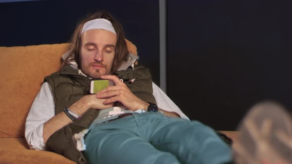 Man with long hair lies on an orange sofa and watches video on a smartphone.