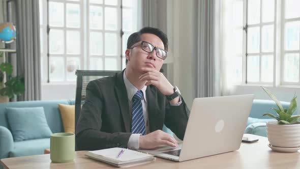 Asian Businessman Thinking And Writing In Notebook While Using Computer For Working At Home alt