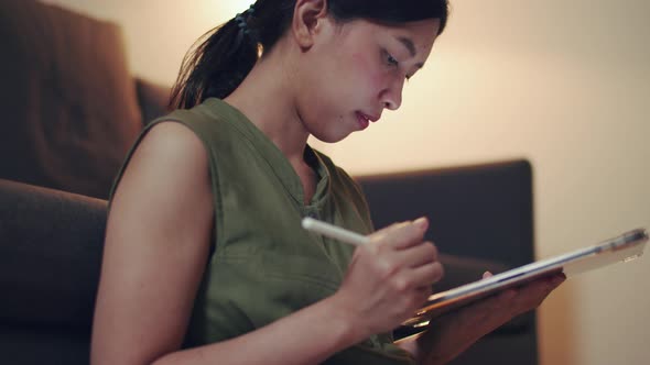 Asian woman reading news on her digital tablet while sitting on the sofa in the living room.