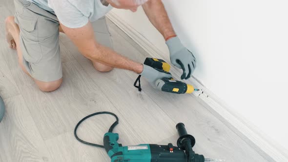 A Man Screws a Skirting Board to the Wall with an Electric Screwdriver in a Room Making Repairs in