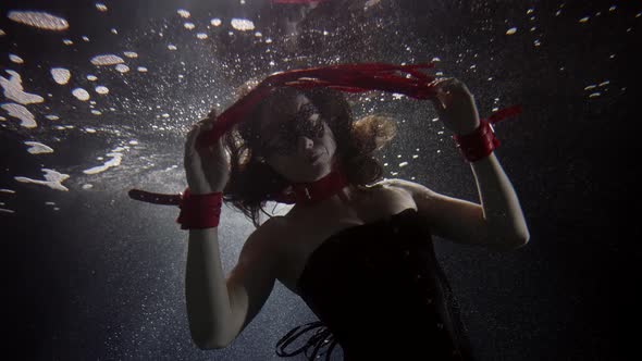 A Darkhaired Woman in Red Handcuffs and with a Red Whip Swims Deep Under the Water on a Dark alt