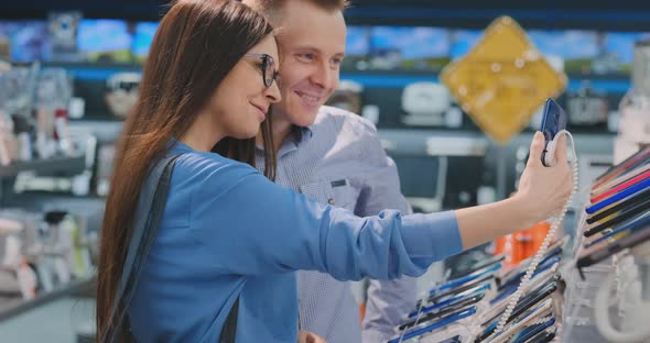 Young Beautiful Married Couple a Man and a Woman Hold a Smartphone Standing Near the Showcase alt
