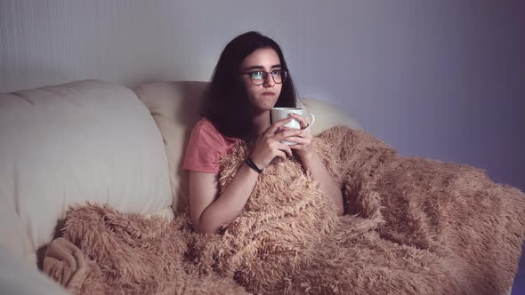 Pretty Girl with a Cup of Tea Sits on the Sofa in the Evening and Watches Tv Glare on Glasses