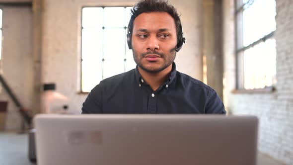 Concentrated Hindu Man Wearing Wireless Headset Using Laptop for Remote Connection alt