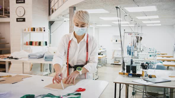 Woman in Protective Mask Tailor of Sewing Workshop is Drawing Pen Around Pattern Laying on Fabric alt