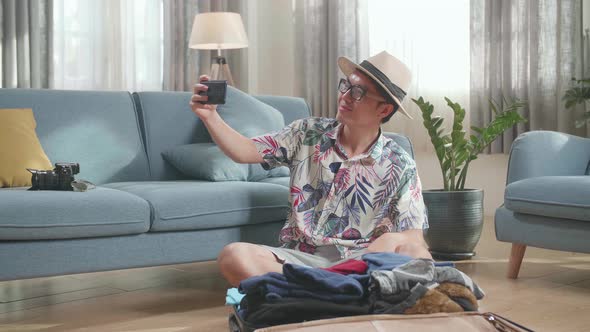 Asian Young Man Try Hat And Selfie While Packing Clothes In Suitcase At Home, Preparing For Vacation alt
