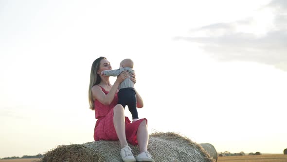 View of Happy Mother Throwing Up and Kissing Her Baby Son on Haystack at Sunset alt