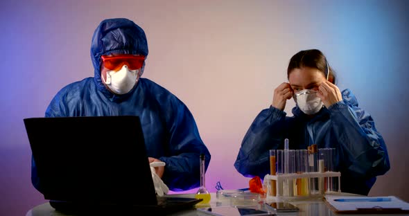 a Man and a Woman in Blue Protective Suits Are Sitting at a Table with a Laptop and Test Tubes. They alt