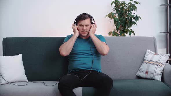 A man sits on the couch wearing headphones on his head, quietly listening to music