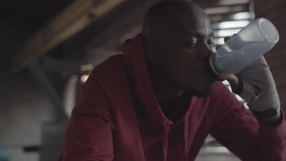 Tired African Boxer in Hoodie Leaning on Ropes and Drinking Water alt