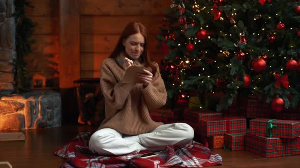 Dreamy Young Woman Writing Christmas Letter To Santa Claus Sitting on Background of Xmas Tree alt
