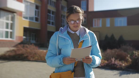 A Teenage Girl in a Blue Jacket and Glasses on the Background of an Educational Institution with a