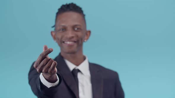 Black Man in an Office Suit Smiling Makes a Gesture of Money with Fingers on a Blue Studio alt