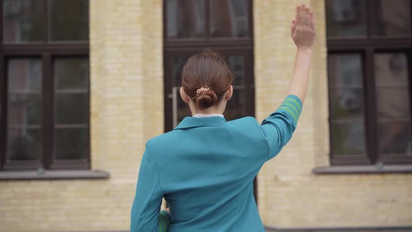 Back View of Slim Brunette Stewardess Standing in Front of Building and Waving. Young Caucasian alt