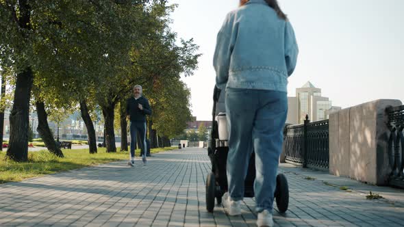 Young Mother Walking with Baby Carriage in City Center While Sportspeople are Running Around alt