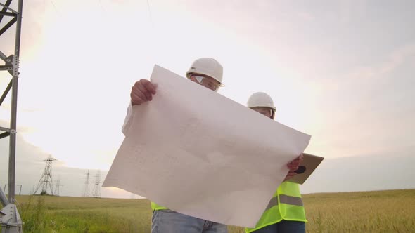 Two Electricians Work Together Standing in the Field Near Electricity Transmission Line in Helmets alt