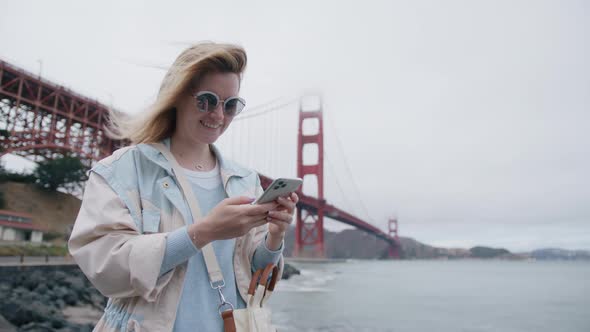 Happy Caucasian Woman Holding Cell Phone Using Smartphone Outdoor at Golden Gate alt