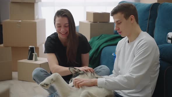 Happy Young Man and Woman Sitting on the Floor in a New Apartment with Cardboard Boxes Laughing and alt