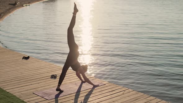 Woman Is Practicing Yoga in Down Facing Dog Pose on One Leg on River Pier.