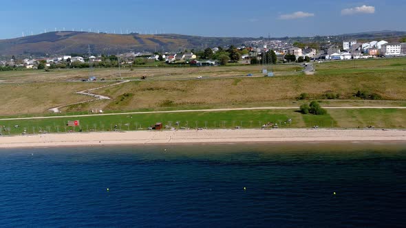 Lake beach with people sunbathing and bathing, garden and walking area, mountains in the background alt