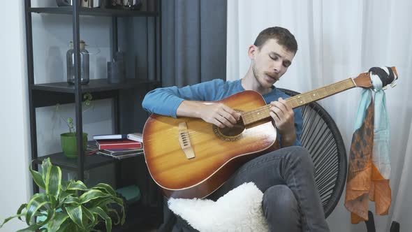 Smiling carefree young man plays acoustic guitar and singing at home in cozy living room. alt