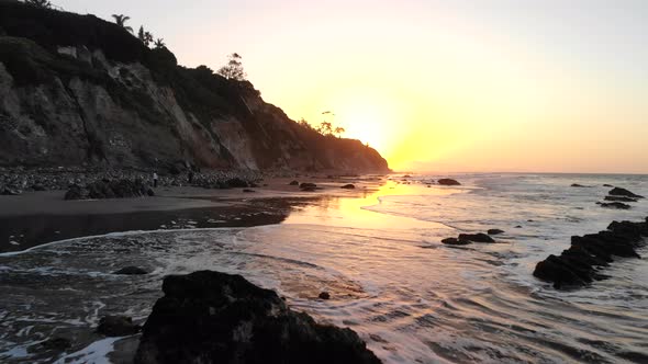 A beach at sunrise with ocean waves and reflections on the shore and cliffs in silhouette along the alt