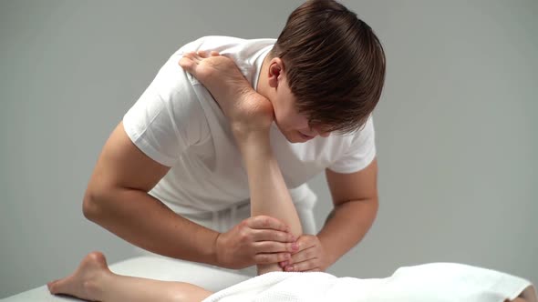 Male Masseur with Strong Hands Massaging Lower Part of Leg to Young Woman Lying on Massage Table alt