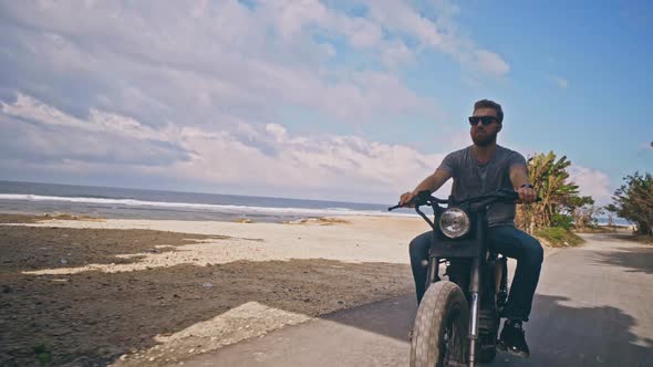 Young Brutal Caucasian Man in Sunglasses Rides Motorcycle on Azure Coast of Bali alt